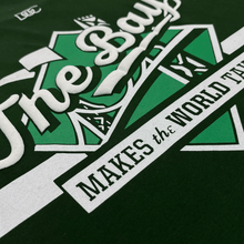 Load image into Gallery viewer, Angled close-up of a white The Bay Makes The World Takes logo graphic on the left chest wearside of a forest green t-shirt.