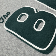 Load image into Gallery viewer, A close-up of gray heavyweight fleece on the Bay Area CC Hoodie By DOC by Oaklandish, featuring bold, textured green and white embroidery spelling "The B"—"The" in cursive and a larger "B"—in men's sizing.
