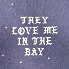 Load image into Gallery viewer, White gothic-style text reading “THEY LOVE ME IN THE BAY” stands out against purple fabric, accented with random white paint splatters. This bold design turns heads and adds a unique flair to any wardrobe—especially when featured on the They Love Me Hoodie By Doc by BY DOC, a heavyweight fleece men’s pullover hoodie designed for both comfort and style.