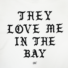 Load image into Gallery viewer, The They Love Me LS Tee By Doc from By DOC features bold, black Gothic-style text in all uppercase letters on a crisp white background, reading “THEY LOVE ME IN THE BAY.” A small By DOC logo with two interlocking C’s is centered beneath the phrase. This simple yet striking design is printed on a heavyweight cotton long sleeve t-shirt for durability and lasting comfort, and is available in men’s sizing.