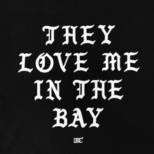 Load image into Gallery viewer, White text on a black background reads “THEY LOVE ME IN THE BAY” in bold, gothic-style uppercase letters. The design appears on the They Love Me Tee By Doc from By DOC’s Bay Collection—a classic fit t-shirt made from heavyweight cotton. A small decorative symbol is centered near the bottom of the artwork.